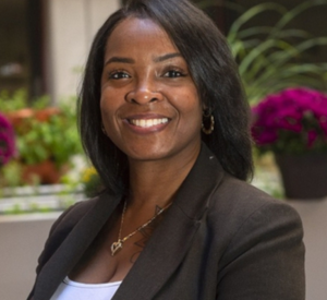 A woman with dark short hair wearing a dark brown blazer over a white shirt with a gold necklace.