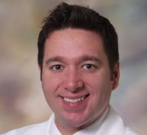 A man with dark brown hair wearing a white collared shirt.