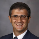 Man with short brown hair and glasses wearing a black suit jacket over a white collared shirt and a black tie.