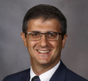 Man with short brown hair and glasses wearing a black suit jacket over a white collared shirt and a black tie.