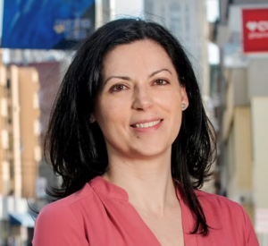 A woman with black hair wearing a pink blouse in front of city buildings.