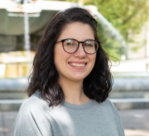 A woman with dark brown hair wearing dark glasses with a gray shirt.