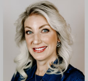 Woman with very light blonde curled hair wearing a dark blue jacket over a black shirt.