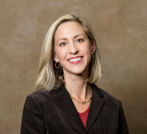 Woman with short blonde hair wearing a brown blazer over a red top and gold earrings.