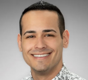 A man with very short black hair wearing a white collared shirt with a black floral pattern.