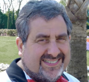 Man with dark gray hair and a beard wearing a white, red and black shirt.