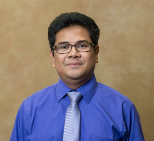 A man with black hair wearing dark-rimmed glasses and a bright blue collared shirt with a silver tie.