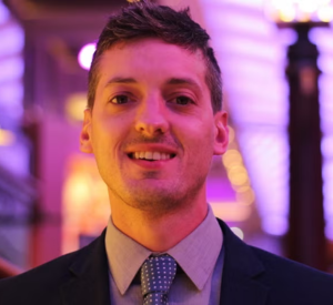 A man with short brown hair wearing a navy blue suit jacker over a tan collared shirt and a gray tie with white polka dots.