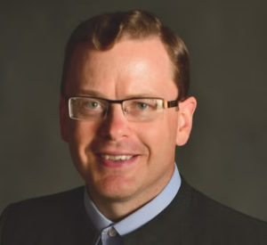 A man with short auburn hair and glasses wearing a black suit jacket over a light blue collared shirt.