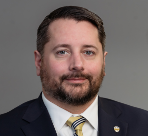 Man with short black hair and a beard wearing a black suit jacket over a white collared shirt with a gold tie with navy blue and white stripes and a University of Pittsburgh pin.