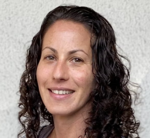 A woman with brown curly hair wearing a dark brown shirt.