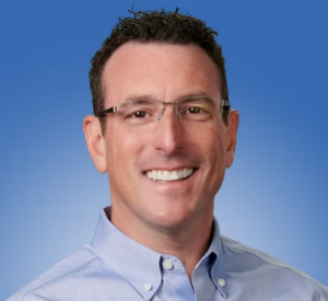 Man with short black hair wearing a light blue collared shirt and glasses.