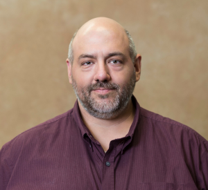 A man with very short gray hair wearing a maroon button-up shirt.