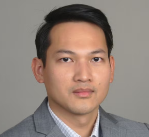 A man with black hair wearing a gray suit jacket over a white collared shirt with gray lines.