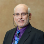 A man with short white hair and glasses wearing a black suit jacket over a purple collared shirt and a blue and purple patterned tie.