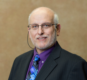 A man with short white hair and glasses wearing a black suit jacket over a purple collared shirt and a blue and purple patterned tie.