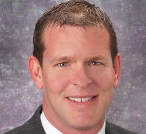 A man with very short brown hair wearing a black suit jacket over a white collared shirt.