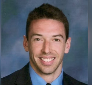 Man with short brown hair wearing a dark suit jacket over a blue collared suit with a dark tie.