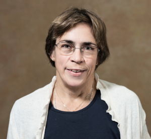 A woman with short brown hair wearing a white cardigan over a black shirt and silver-rimmed glasses.