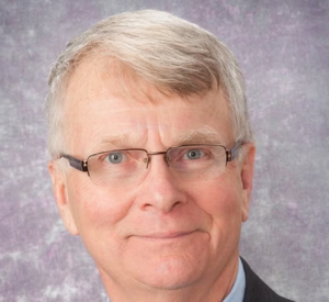 Man with white hair wearing wire glasses and a black suit jacket over a light blue collared shirt.