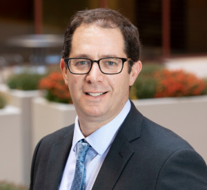 A man with brown hair and dark-rimmed glasses wearing a dark gray suit jacket over a white collared shirt and a blue tie.