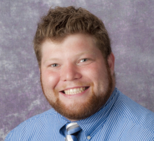 Man with short auburn hair and beard wearing a blue and white striped collared shirt with a blue and white tie.