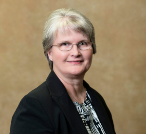 A woman with short white hair with silver-rimmed glasses wearing a black cardigan over a black and white blouse.