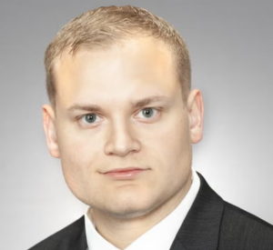 Man with short blonde hair wearing a black suit jacket over a white collared shirt.