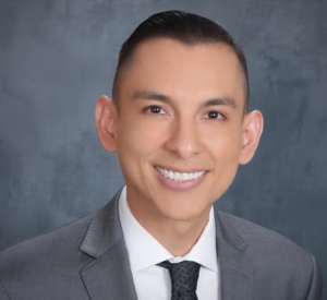 A man with very short black hair wearing a gray suit jacket over a white collared shirt and a black tie.