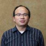 A man with short black hair and black-rimmed glasses wearing a navy blue collared shirt with white stripes.