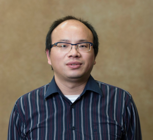 A man with short black hair and black-rimmed glasses wearing a navy blue collared shirt with white stripes.