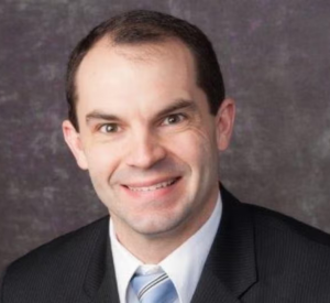 A man with very short dark hair wearing a black suit jacket over a white collared shirt and a light blue tie.