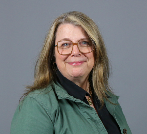 Woman with blonde hair wearing light brow glasses and a green collared shirt over a black blouse.