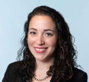 A woman with dark brown curly hair wearing a black blazer and a silver beaded neckalce.