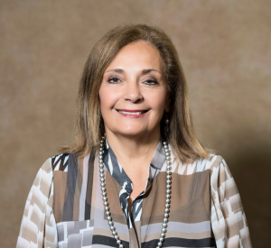 A woman with short brown hair wearing a dark and light brown patterned blouse with a silver beaded necklace.