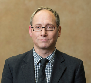 A man with short blonde hair wearing a black suit jacket over a black and white checkered collared shirt with a black tie.