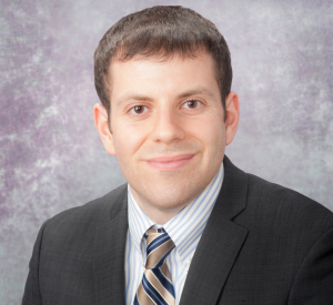 A man with short brown hair wearing a black suit jacket over a white and gray striped collared shirt with a gold and blue striped tie.