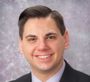 Man with brown hair wearing a black suit jacket over a light gray collared shirt.