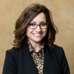 A woman with brown hair and black-rimmed glasses wearing a black blazer over a collared shirt with a black and brown pattern.