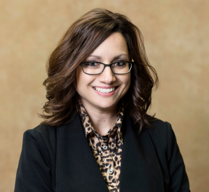 A woman with brown hair and black-rimmed glasses wearing a black blazer over a collared shirt with a black and brown pattern.