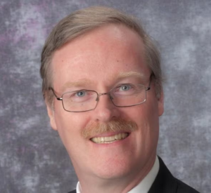 Man with short blonde hair and a mustache wearing wired glasses and a black suit jacket over a white collared shirt.