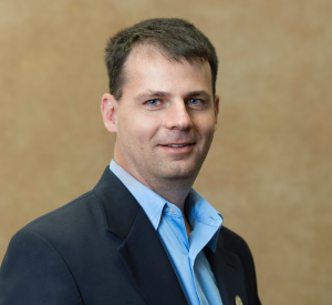 A man with short brown hair waring a navy blue suit jacket over a light blue collared shirt.