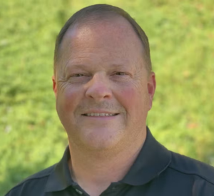 A man with very short auburn hair wearing a black collared shirt.