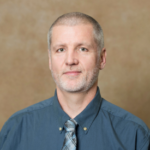 A man with short gray hair wearing a teal collared shirt and a gray and teal striped tie.