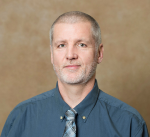 A man with short gray hair wearing a teal collared shirt and a gray and teal striped tie.