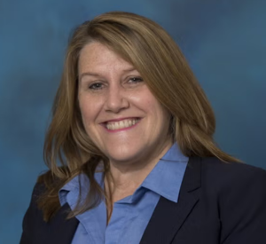 Woman with dark blonde hair wearing a black blazer over a blue collared shirt.