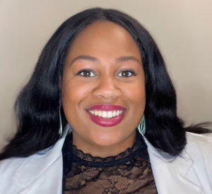 A woman with black hair wearing a white lab coat over a black lace shirt.