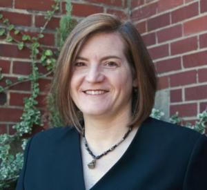 Woman with short auburn hair wearing a black blazer and a black and brown beaded necklace.