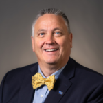 A man with short gray hair wearing a black suit jacket over a blue collared shirt and a yellow bowtie.