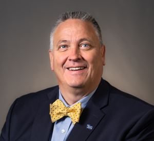 A man with short gray hair wearing a black suit jacket over a blue collared shirt and a yellow bowtie.